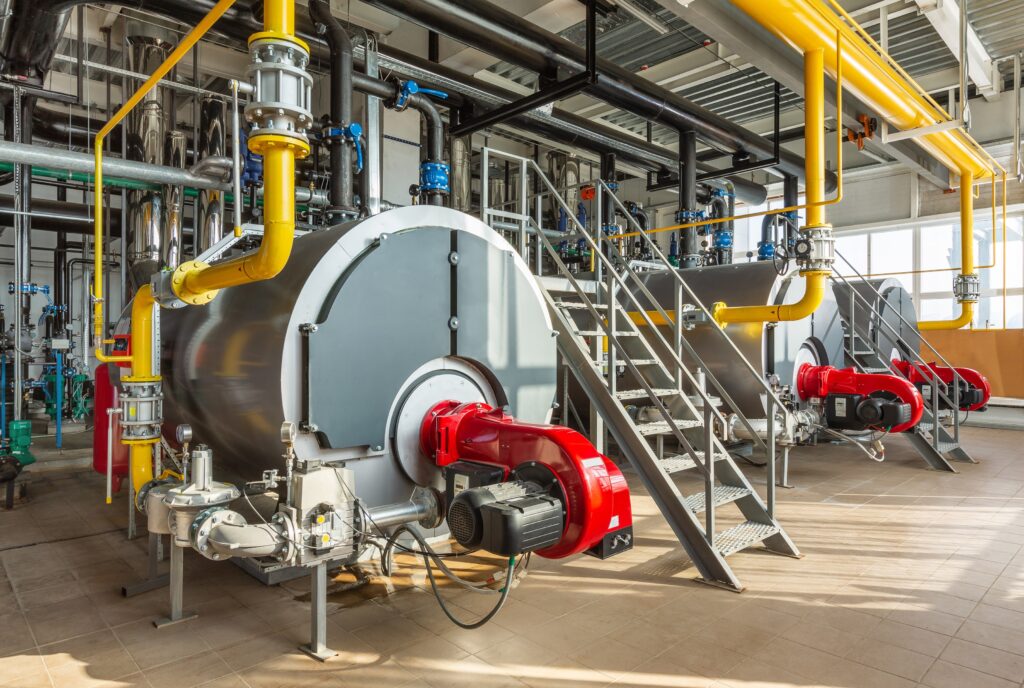 Industrial steam boilers with bright yellow gas piping and red blowers, used in high-pressure systems vulnerable to boiler carryover.