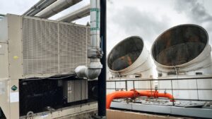 A cooling tower vs chiller comparison, showcasing the differences between these cooling systems. On the right, a cooling tower, often used in power plants and industrial processes, utilizes evaporative cooling to remove excess heat and reject heat into the surrounding air. On the left, an industrial chiller (air-cooled) provides efficient cooling in large-scale HVAC systems, such as data centers and commercial buildings, through a refrigeration cycle. Both systems, including water-cooled chillers and air-cooled chillers, are critical for precise temperature control, cooling needs, and maximizing energy efficiency in industrial applications, where heat exchangers and evaporative cooling play vital roles in dissipating heat.